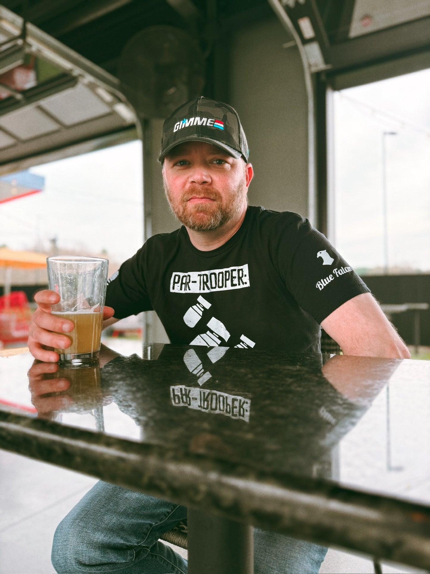 A man at the bar with a drink in his hand wearing the Blue Falcon "Gimmie" hat and the Blue Falcon "Par-trooper" t-shirt.