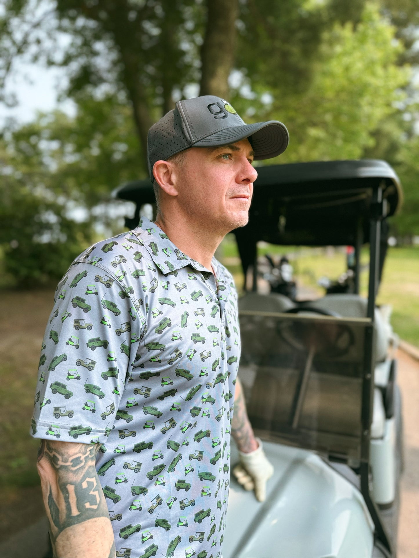 A man wearing a Blue Falcon "Motor Tee" golf polo featuring a playful all-over print of Jeeps, Humvees, and golf carts. He is also wearing a Blue Falcon grey "Birdie Bomber" hat.
