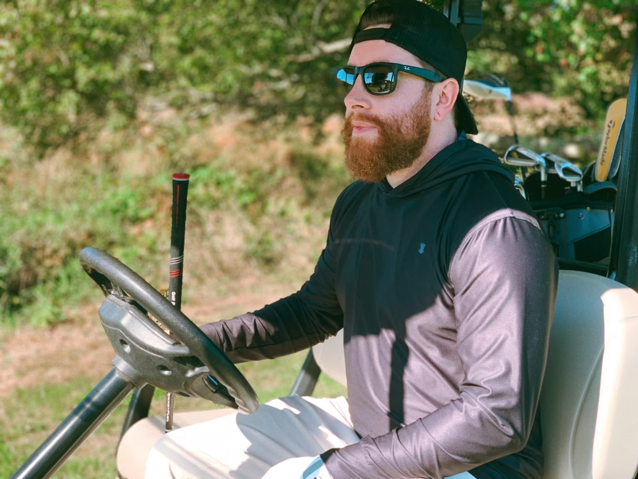 A man sitting in a golf cart wearing a gray and black trucker hat with a falcon logo and "Blue Falcon" written on the side, paired with a performance hoodie featuring icons inside the hood.