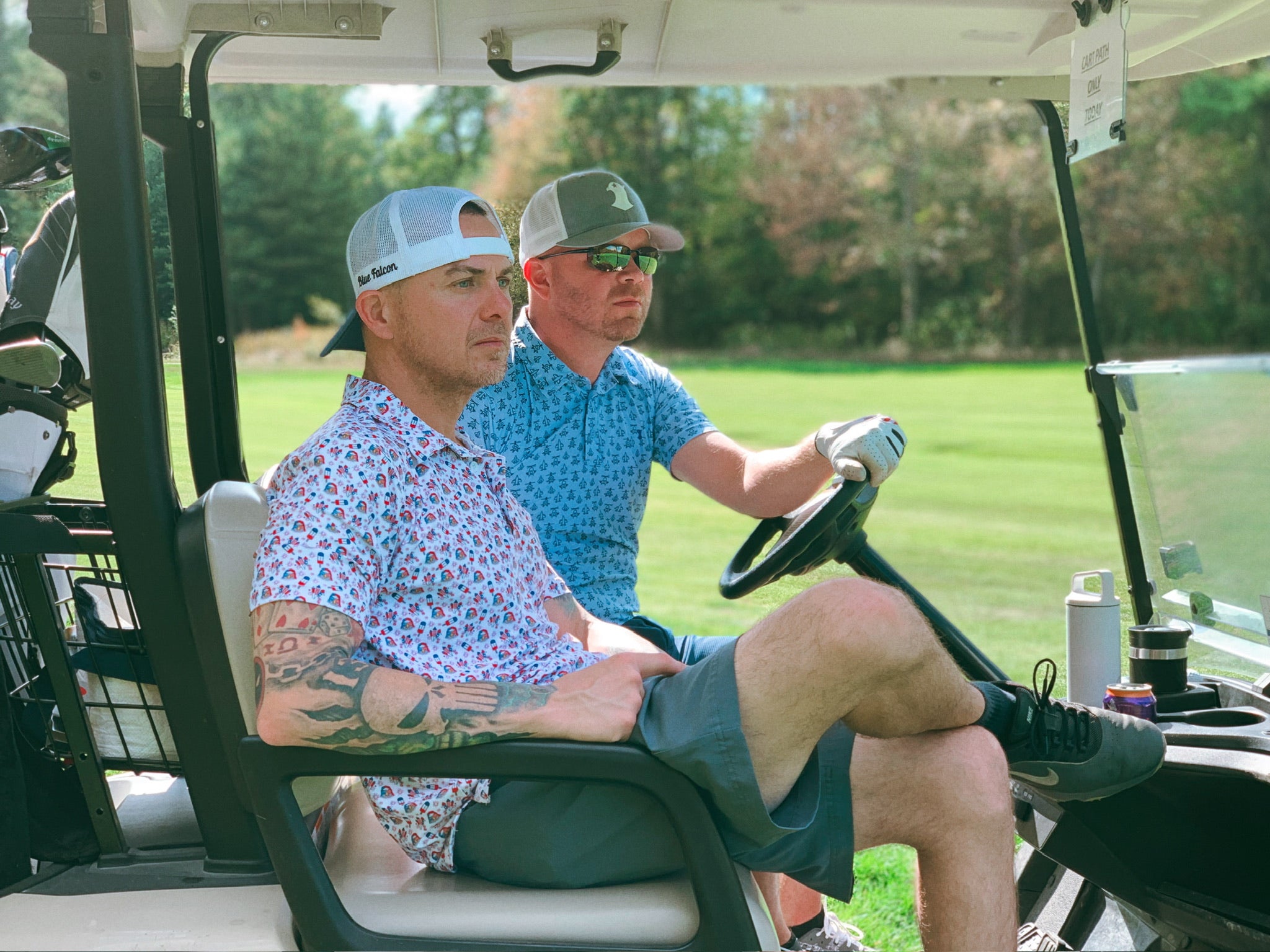 Two men cruising in a golf cart wearing stylish performance polos and golf hats, scanning the surroundings.