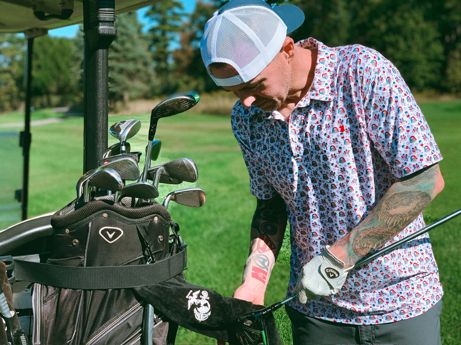 Golfer wearing a red white and blue party eagle polo and blue and blue falcon  white trucker hat cleaning his golf club at his cart