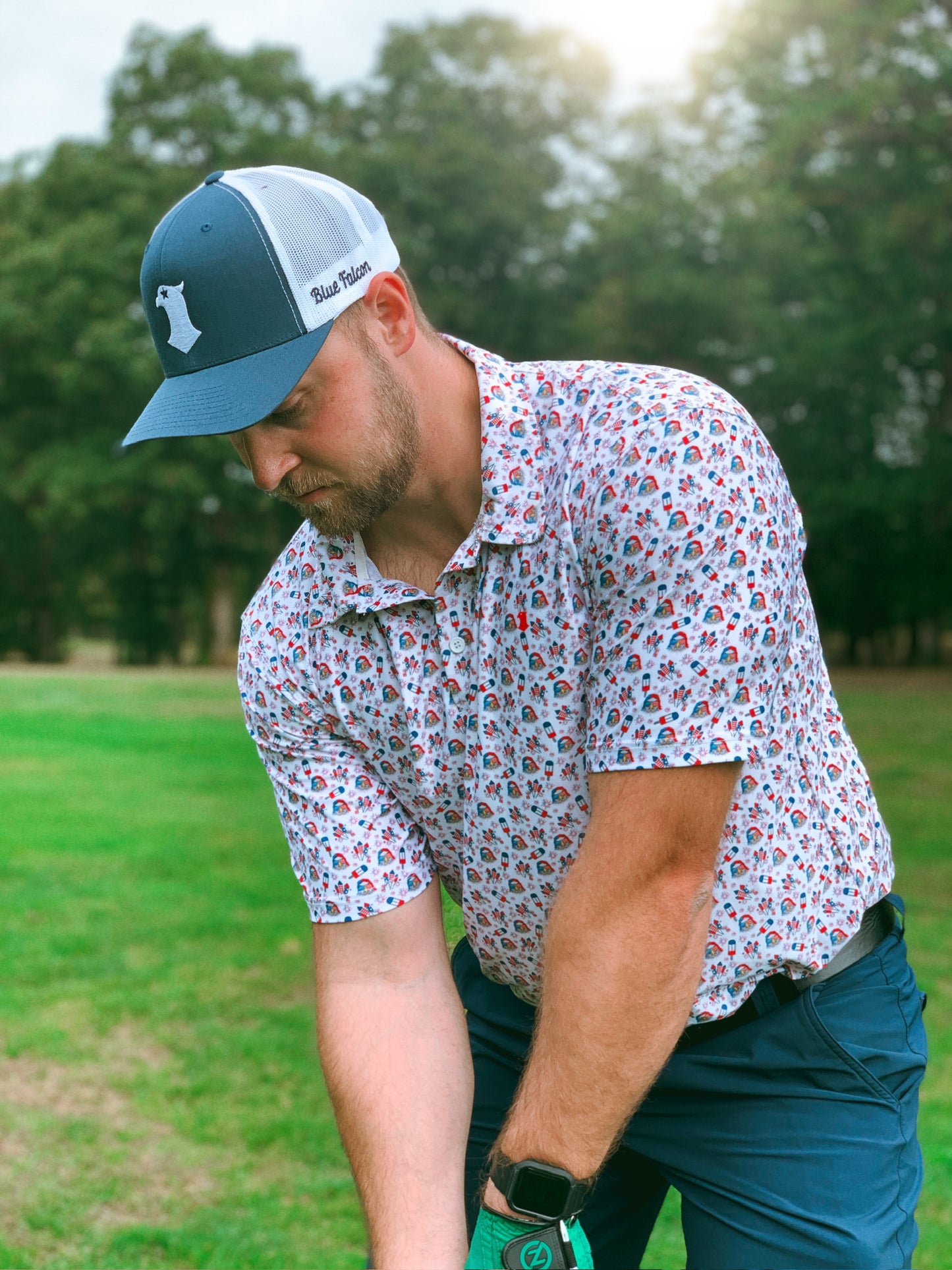 A golfer in a white performance polo with patriotic icons, and a blue and white trucker hat with a falcon logo and "Blue Falcon" written on the side, setting up to the ball.