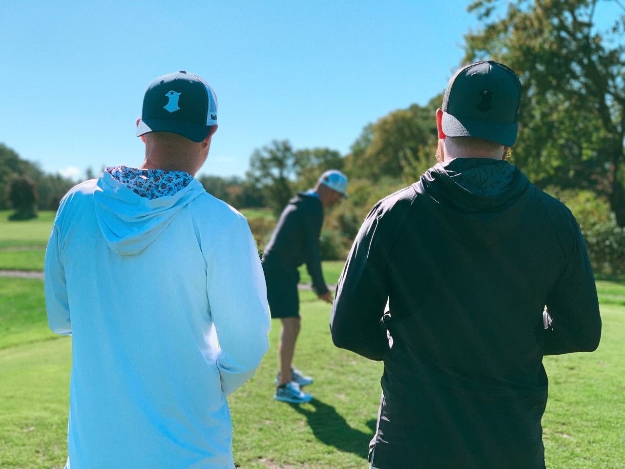 Three friends on a golf course, two watching as one prepares to swing. They are all wearing blue falcon golf hats and performance hoodies with graphic designs inside the hood.