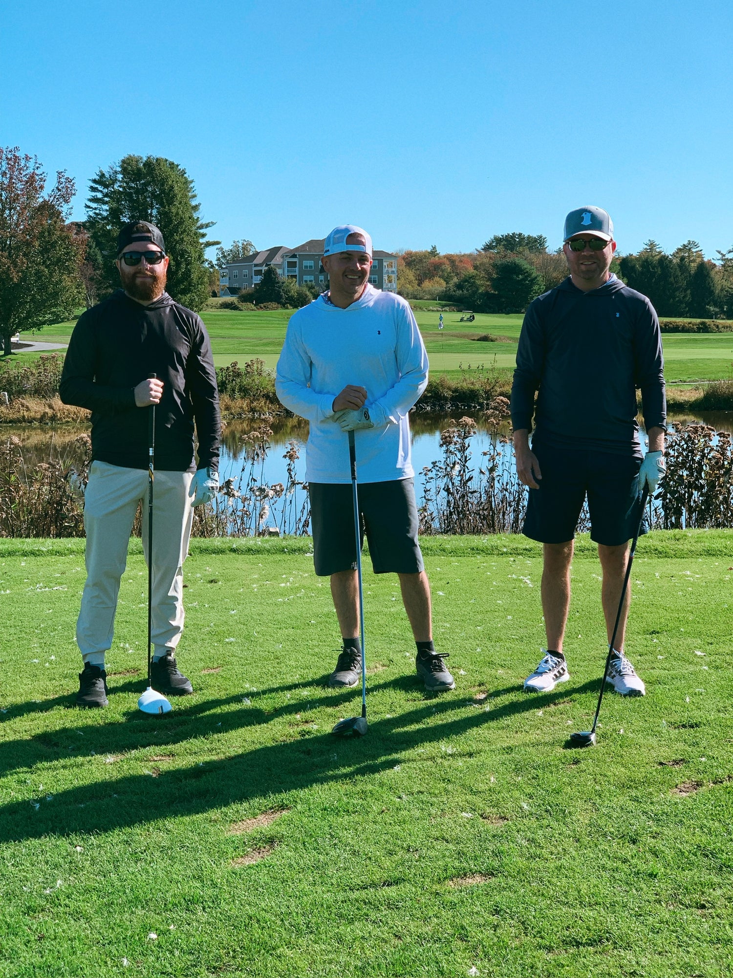 Three men in golf attire, wearing golf hats and performance hoodies, stand together on the golf course holding golf clubs. They are looking straight ahead, clearly focused and ready to take their swings.