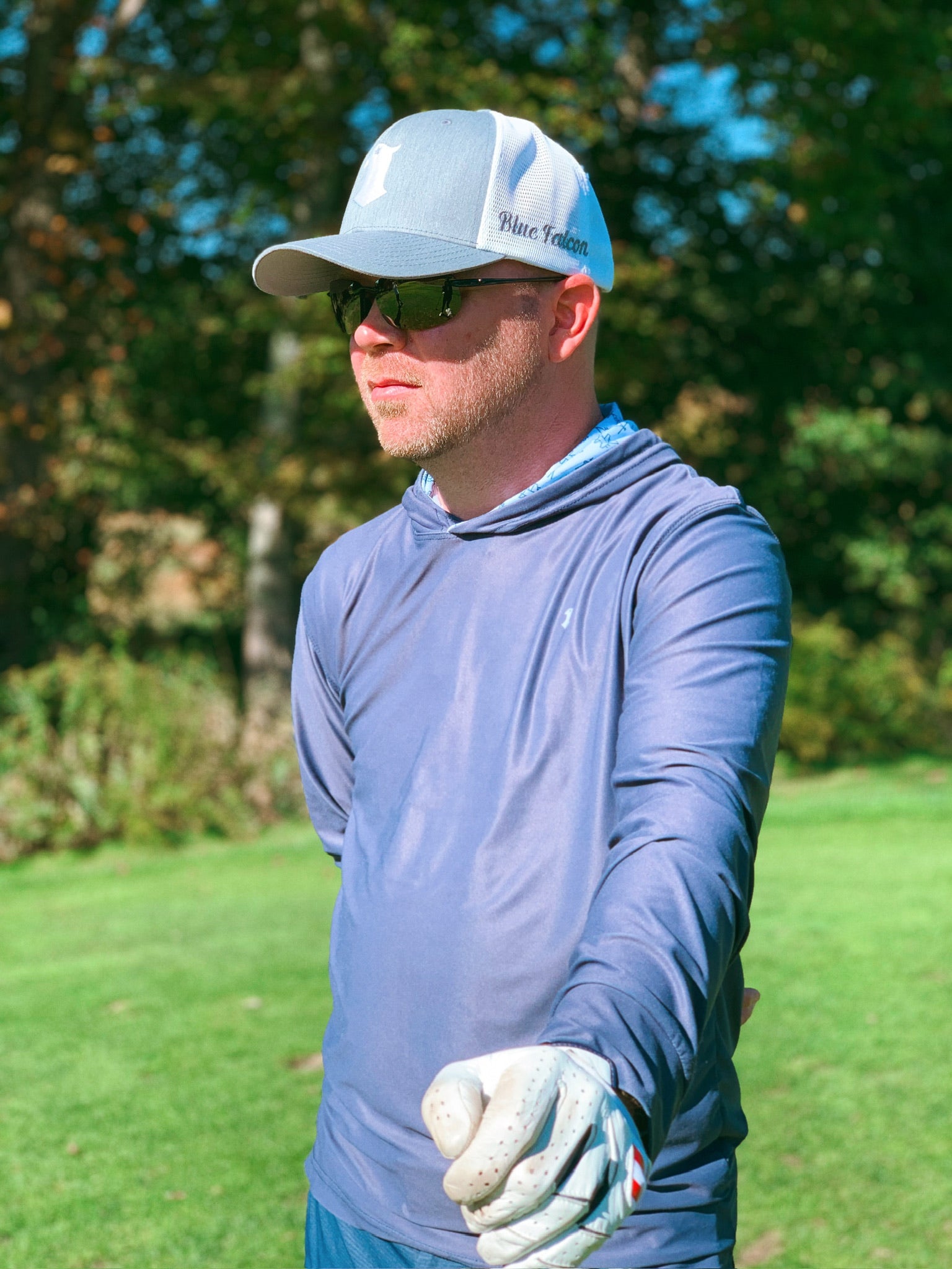 A golfer wearing a navy blue performance hoodie with graphic print inside the hood, a gray hat with a falcon logo on the front and "Blue Falcon" written on the side, looking off into the distance.