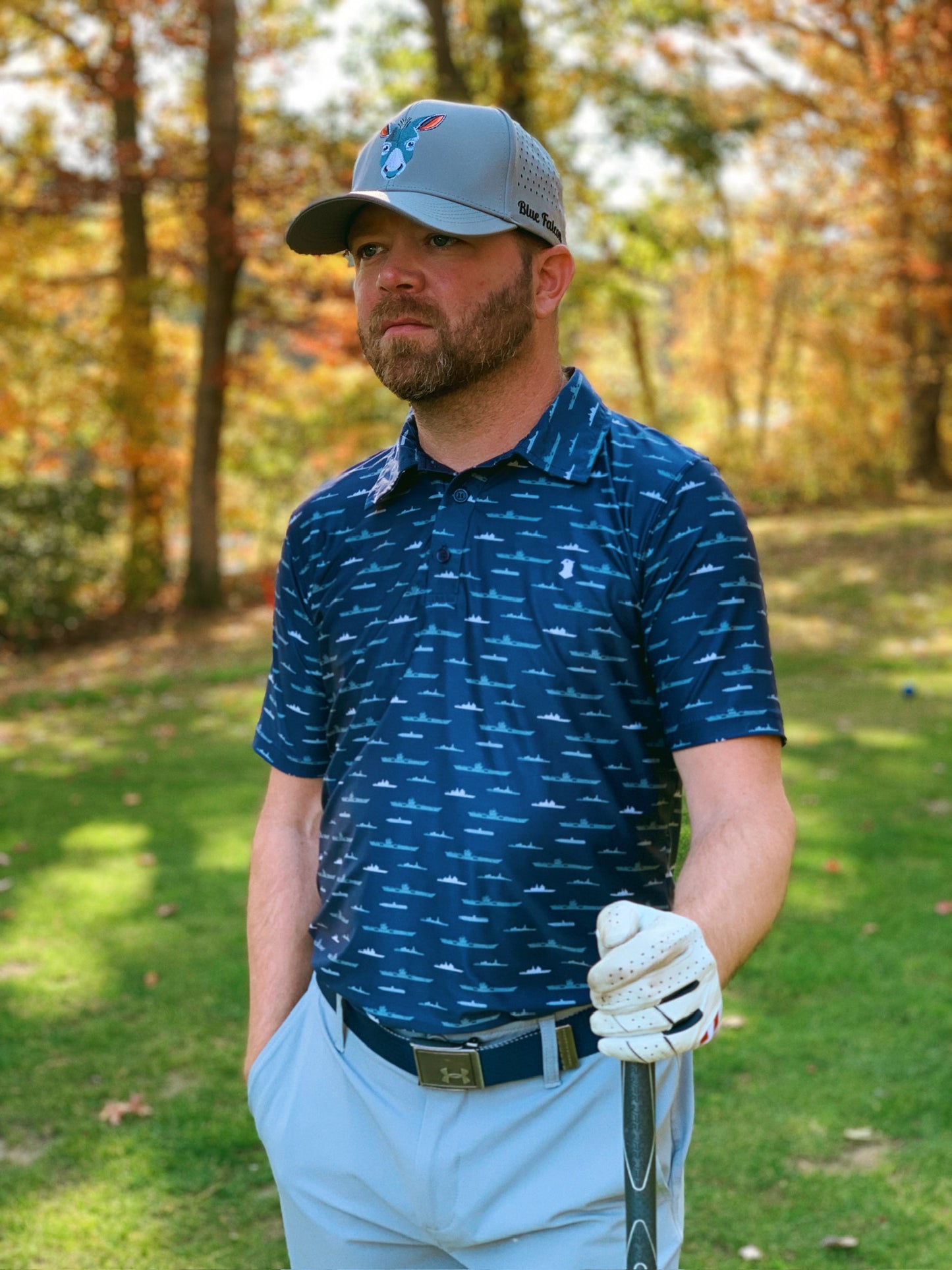 A golfer wearing a navy blue performance polo with ships and submarines print, a gray hat with a donkey on the front and "Blue Falcon" written on the side, looking off into the distance.