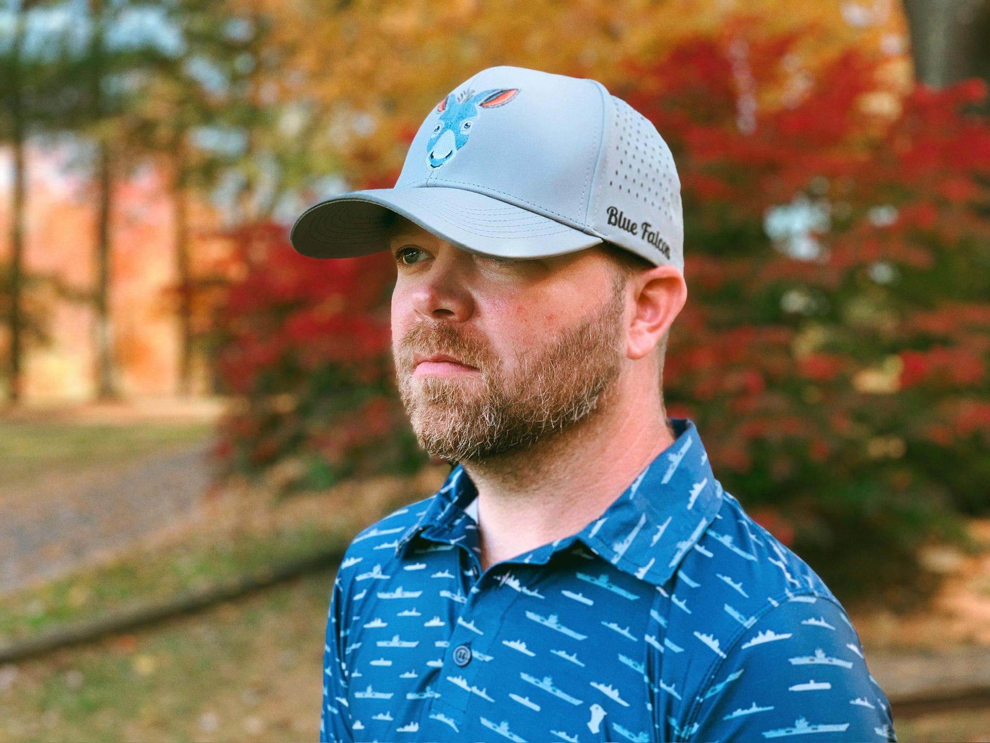 A golfer wearing a navy blue performance polo with ships and submarines print, a gray hat with a donkey on the front and "Blue Falcon" written on the side, looking off into the distance.