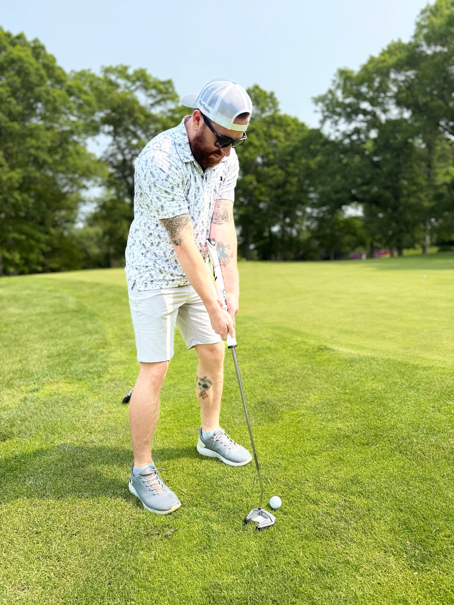 A man wearing a Blue Falcon golf polo featuring an all-over print of pineapple grenades and crossed golf clubs. He is putting from the fringe. 